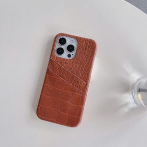 Puede incluir: Una funda para smartphone de color &oacute;xido con un patr&oacute;n texturizado de piel de cocodrilo. La parte posterior de la funda incluye una ranura diagonal para tarjetas, con una textura de grano m&aacute;s fino en la parte superior y un patr&oacute;n de cuadros grandes.