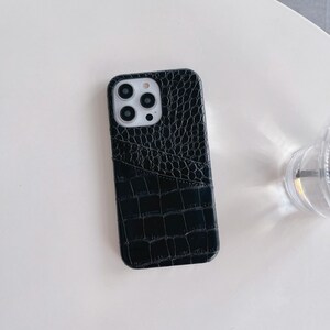 Puede incluir: Una funda para smartphone de color negro con una textura brillante de piel de cocodrilo. La funda incluye un bolsillo para tarjetas integrado en la parte posterior y un recorte para las lentes de la c&aacute;mara, dispuestas en un m&oacute;dulo cuadrado.