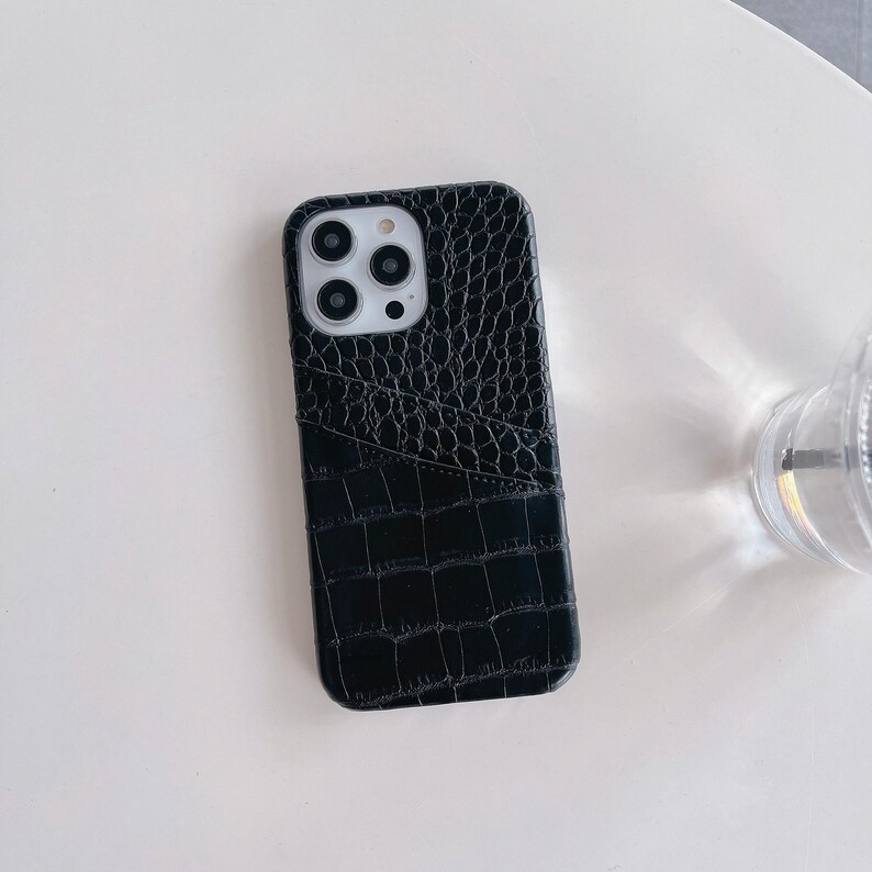 Puede incluir: Una funda para smartphone de color negro con una textura brillante de piel de cocodrilo. La funda incluye un bolsillo para tarjetas integrado en la parte posterior y un recorte para las lentes de la c&aacute;mara, dispuestas en un m&oacute;dulo cuadrado.