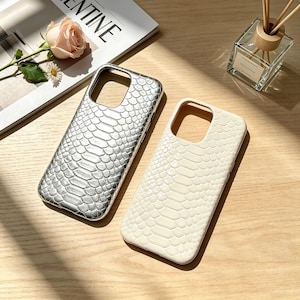Peut inclure: Deux coques de smartphone rectangulaires présentant un motif texturé en peau de serpent. L'une est finie dans une couleur argentée métallique, tandis que l'autre est d'une couleur blanc cassé unie. Les deux ont une découpe pour l'objectif.