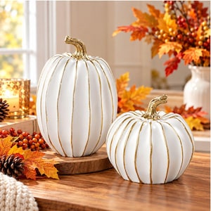 May include: Two white decorative pumpkins with vertical gold-painted ridges and gold stems. The larger pumpkin is tall and oval-shaped, while the smaller pumpkin is shorter and rounder. Both feature a smooth finish and metallic gold accents.