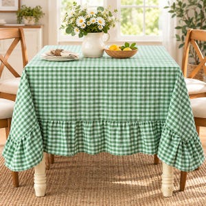 May include: A square tablecloth featuring a green and white gingham check pattern with a ruffled edge. The tablecloth is draped over a table, topped with a white ceramic pitcher filled with white daisies and a bowl of yellow lemons.