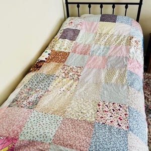 May include: A single-sized patchwork quilt featuring a grid of square fabric patches in various floral, striped, and solid pastel patterns including pink, blue, yellow, and white. The quilt is draped over a bed with a black metal headboard frame.