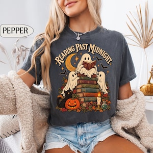 May include: A charcoal gray t-shirt featuring a graphic print with the text "READING PAST MIDNIGHT" above a stack of books, three ghosts, a black cat, a jack-o'-lantern, a crescent moon, bats, a lit candle, and a small potion bottle.