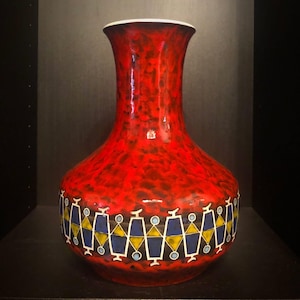 Può includere: Un vaso in ceramica alto caratterizzato da una vivace smaltatura rossa maculata sul collo e sulla parte superiore. La sezione inferiore è decorata con un motivo geometrico ripetitivo di forme blu e gialle bordate di bianco su fondo scuro.