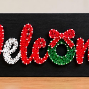 Puede incluir: Un letrero rectangular de madera negra con la palabra Welcome en arte con hilos. Las letras están creadas con hilo rojo y blanco y clavos pequeños. Una hoja de acebo de hilo verde está en cada extremo y una corona reemplaza la letra o.