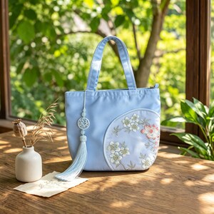 May include: A light blue handbag featuring two top handles and a circular side panel adorned with white and peach-colored floral embroidery. A decorative light blue tassel with a white circular charm hangs from the side of the bag.