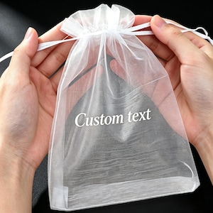 Può includere: Un sacchetto con coulisse in organza bianca traslucida con il testo Custom text stampato in bianco sul davanti. La borsa è realizzata in un tessuto leggero e trasparente con una chiusura a nastro bianco abbinata nella parte superiore.