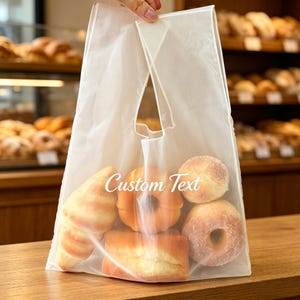 Könnte beinhalten: Eine halbtransparente weiße Einkaufstasche mit integrierten Griffen, die verschiedene Backwaren wie ein Croissant, einen Krapfen und Brötchen enthält. Auf der Vorderseite der Tasche ist der weiße kursive Schriftzug Custom Text aufgedruckt.