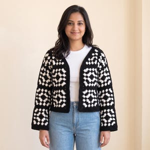 May include: A long-sleeved open-front cardigan constructed from black and white crocheted granny squares. The garment features a repeating geometric pattern, a V-neckline, and a straight hem, worn over a plain white t-shirt and light blue jeans.