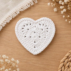 May include: A white heart-shaped crochet coaster featuring a textured, stitched pattern with a braided border edge. The piece is crafted from white yarn and displays a symmetrical design with small, evenly spaced openings throughout the surface.