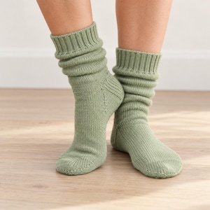 May include: A pair of sage green knit socks featuring a ribbed cuff and a slouchy, mid-calf length design. The socks are constructed with a standard knit pattern, including a reinforced heel section and a smooth, rounded toe area.