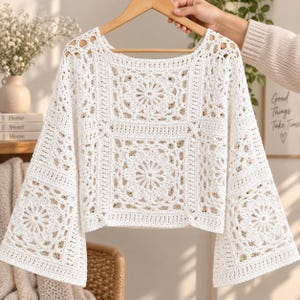 May include: A white long-sleeve top crafted from crochet fabric featuring a square neckline and a repeating floral granny square pattern. The garment has wide, flared sleeves and a cropped length, constructed with intricate yarn stitching throughout.