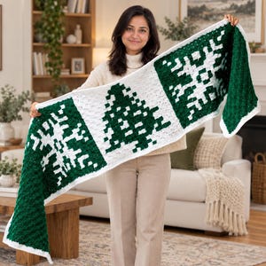 May include: A rectangular crochet scarf featuring a repeating pattern of white snowflakes and a central green pine tree on a dark green background. The scarf is finished with a white border and has a textured, yarn-based construction.