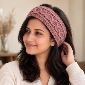 May include: A dusty rose crochet headband featuring a prominent central cable knit pattern bordered by ribbed edges. The accessory is crafted from yarn with a textured finish, designed to sit across the forehead and crown of the head.