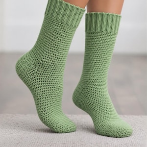 May include: A pair of sage green mid-calf socks crafted with a dense crochet stitch pattern. The socks feature a ribbed cuff at the top, a reinforced heel section, and a rounded toe, providing a consistent texture throughout the entire garment.