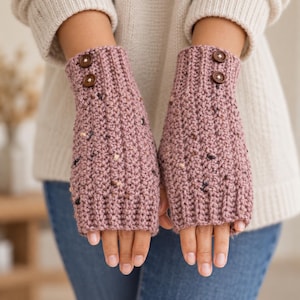 May include: A pair of mauve fingerless gloves featuring a textured knit pattern with small dark and light flecks throughout. Each glove is accented with two round brown buttons positioned near the top edge, providing a decorative detail.