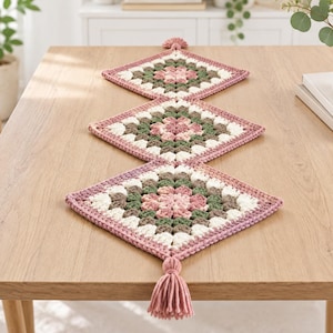 Puede incluir: Un camino de mesa de ganchillo con tres motivos cuadrados de la abuela en forma de diamante en tonos rosa empolvado, verde salvia y crema. El camino está rematado con una borla de hilo rosa en cada extremo, con una textura artesanal.
