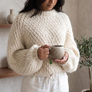 May include: A cream-colored, chunky knit sweater with a ribbed crew neckline and long, voluminous sleeves. The sweater features a textured, raised stitch pattern throughout. It is paired with white drawstring pants and a speckled ceramic mug.