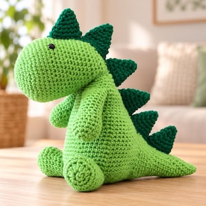 Peut inclure: Un jouet dinosaure crocheté à la main de couleur vert citron vif. La créature présente une tête arrondie, de petits bras et une longue queue. Une rangée de pointes triangulaires vert forêt foncé longe le dos et le sommet de la tête. Yeux en perles noires.
