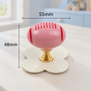 May include: A pink ceramic cabinet knob shaped like an American football with red laces, mounted on a gold-toned metal base and a white four-lobed plate. The knob measures 55mm in width and 48mm in height as indicated by the visible labels.