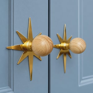 May include: Two cabinet knobs featuring a natural light wood spherical handle attached to a polished gold-tone metal starburst backplate. The star-shaped hardware is mounted onto a matte blue cabinet door with vertical panel detailing.