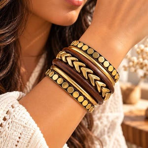May include: A stack of five bracelets featuring two black bands with gold-toned circular studs, two smooth gold-toned metal bands, and one dark brown wooden band with a central row of gold-toned chevron-shaped metal accents.
