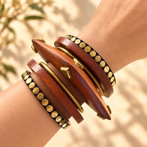 Puede incluir: Un conjunto de pulseras multicapa que presenta dos bandas negras delgadas adornadas con pequeños tachones circulares dorados, dos bandas de madera marrón oscuro lisa y una pieza central de madera gruesa y curva con herrajes dorados.