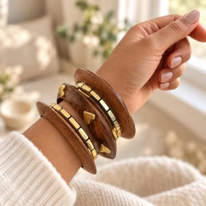 Op de afbeelding: Een set van drie gestapelde armbanden met donkerbruine, gepolijste houten banden, geaccentueerd met metallic goudkleurige rechthoekige segmenten en kleine goudkleurige hartvormige bedels. De armbanden hebben een glad, gebogen profiel en glans.