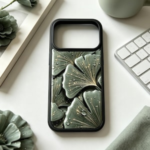Op de afbeelding: Een zwarte smartphonehoes met een patroon van saliegroene ginkgobladeren met gouden nerven en kleine waterdruppels. Het ontwerp bedekt de achterkant van de hoes, die aan de bovenkant een rechthoekige uitsparing voor de camera heeft.