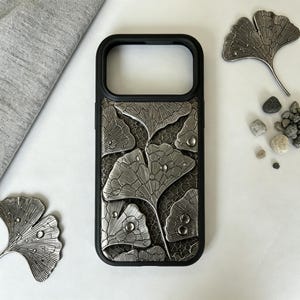 Op de afbeelding: Een zwarte telefoonhoes met een metallic zilveren reliëfontwerp van overlappende ginkgobladeren met kleine doorzichtige druppels op het oppervlak. De achtergrond van de hoes heeft een fijn, donker en ingewikkeld gaaspatroon.