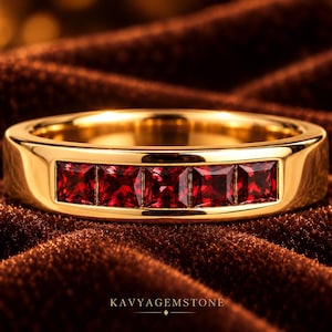 May include: A polished gold band ring featuring five square-cut, deep red gemstones set in a horizontal channel. The metal surface is smooth and reflective, showcasing a bright yellow gold tone. The text KAVYAGEMSTONE is printed at the bottom.