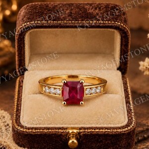 May include: A gold ring featuring a rectangular, deep red gemstone at the center, held by four prongs. The band is accented with three small, round, clear stones set into each side of the shank, leading up to the central red stone.