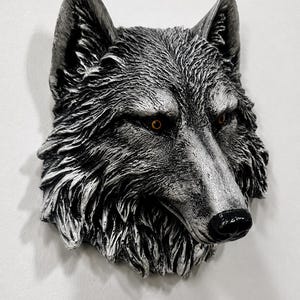 May include: A three-dimensional wall sculpture of a wolf head featuring detailed fur textures in shades of silver, grey, and black. The wolf has pointed ears, a black nose, and piercing amber-coloured eyes that stand out against the monochromatic fur.