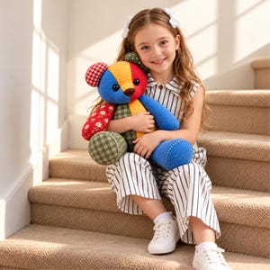 May include: A plush teddy bear constructed from various fabric patches, including red and white gingham, yellow polka dots, red floral print, and solid blue and green textures. The bear features dark round eyes and a brown stitched nose.