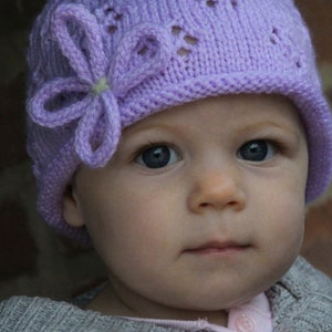 May include: A close-up of a baby wearing a purple knitted hat with a flower design. The hat has a delicate crocheted pattern and a soft, feminine look.
