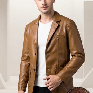 Peut inclure: Un blazer en cuir marron clair avec un revers cranté, deux boutons foncés, une poche poitrine et deux poches plaquées inférieures. La veste est portée sur un t-shirt blanc uni. Un écusson rond en cuir marron avec le texte HC HIDE & CRAFT est visible.