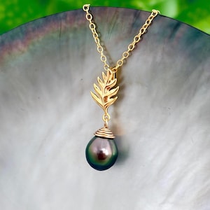 May include: A gold-colored necklace featuring a leaf-shaped pendant and a large, dark pearl. The pearl has a deep, iridescent color with hints of green and purple. The necklace is displayed against a neutral background.