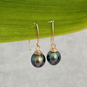 Puede incluir: Un par de pendientes colgantes dorados con perlas de color gris verdoso oscuro. Las perlas redondas tienen un brillo metálico. Los pendientes tienen un diseño en espiral encima de la perla y un gancho simple.