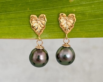 May include: A pair of gold-colored leaf-shaped earrings with dark gray, round pearls. The earrings are hanging against a green leaf background. The leaf-shaped tops are detailed with vein patterns.