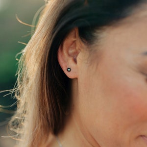 May include: A close-up of a person's ear with a small, dark green pearl earring. The earring is a stud style and is set in a silver setting.