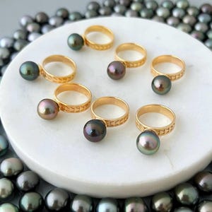 Pode incluir: Anéis de ouro com um design geométrico, cada um com uma grande pérola iridescente e escura. Os anéis estão dispostos sobre uma superfície branca, com um colar de pérolas no fundo.