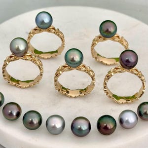 May include: Gold rings with floral designs, each topped with a large, lustrous pearl in shades of green, gray, and purple. A collection of loose pearls in similar colors are arranged below the rings. The jewelry is displayed on a white surface.