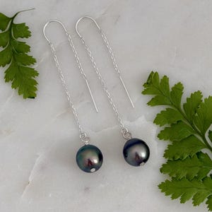 Puede incluir: Un par de pendientes de hilo de plata con perlas grises oscuras e iridiscentes. Los pendientes presentan una delicada cadena y un diseño de gancho curvo. Las perlas son redondas y tienen un brillo sutil. Fondo con hojas de helecho verdes.