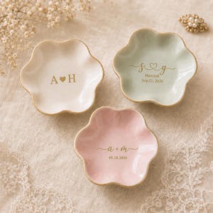 May include: Three flower-shaped ceramic trinket dishes in cream, sage green, and soft pink with gold-painted edges. Each dish features gold-printed personalization: A H with a heart, S g with a heart and date, and a + m with a date.