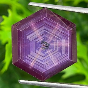 May include: A hexagonal-cut purple gemstone featuring a distinct star-like pattern with concentric geometric lines radiating from the center. The stone is held by silver-toned metal tweezers, showcasing its deep violet hue and symmetrical facets.