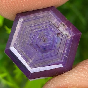 Könnte beinhalten: Ein sechseckiger violetter Edelstein mit einem ausgeprägten geometrischen Muster aus konzentrischen Ringen, die von der Mitte ausstrahlen. Der Stein hat eine glatte, polierte Oberfläche mit sichtbarer innerer Bänderung und einem tiefvioletten Farbton.