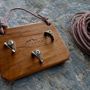 May include: A rectangular wooden climbing hold board with three metal climbing holds attached to the front. The board features the text LITTLE ROCK with a mountain graphic. A red and black patterned rope is threaded through two top holes.