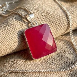 May include: A silver chain necklace featuring a square-shaped, faceted, deep pink gemstone pendant. The pendant is set in a simple silver frame with a small bail connecting it to the delicate, thin silver link chain that rests in a circular shape.