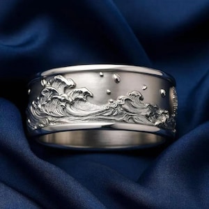 May include: A wide silver band ring featuring a detailed, raised relief carving of crashing ocean waves across the outer surface. The metal has a brushed, matte finish, and the design includes small, circular accents resembling sea spray.